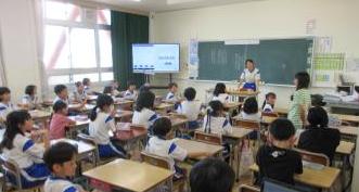 教室で机に座った児童たちが教壇に立つ児童と立って発表する児童を見ている様子